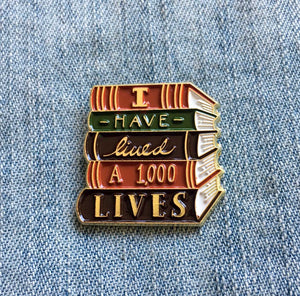 By Ectogasm. The I Have Lived 1,000 Lives Pin is a gorgeous enamel pin for book lovers. It features a stack of books with the quote, "I Have Lived 1,000 Lives". This durable pin is made with gold plated metal and enamel. It is kept secure with a metal clasp. Use it to show your love of reading with every outfit. Makes a charming gift for readers and librarians.  Measures 1" x 1"