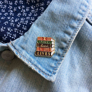 By Ectogasm. The I Have Lived 1,000 Lives Pin is a gorgeous enamel pin for book lovers. It features a stack of books with the quote, "I Have Lived 1,000 Lives". This durable pin is made with gold plated metal and enamel. It is kept secure with a metal clasp. Use it to show your love of reading with every outfit. Makes a charming gift for readers and librarians.  Measures 1" x 1"